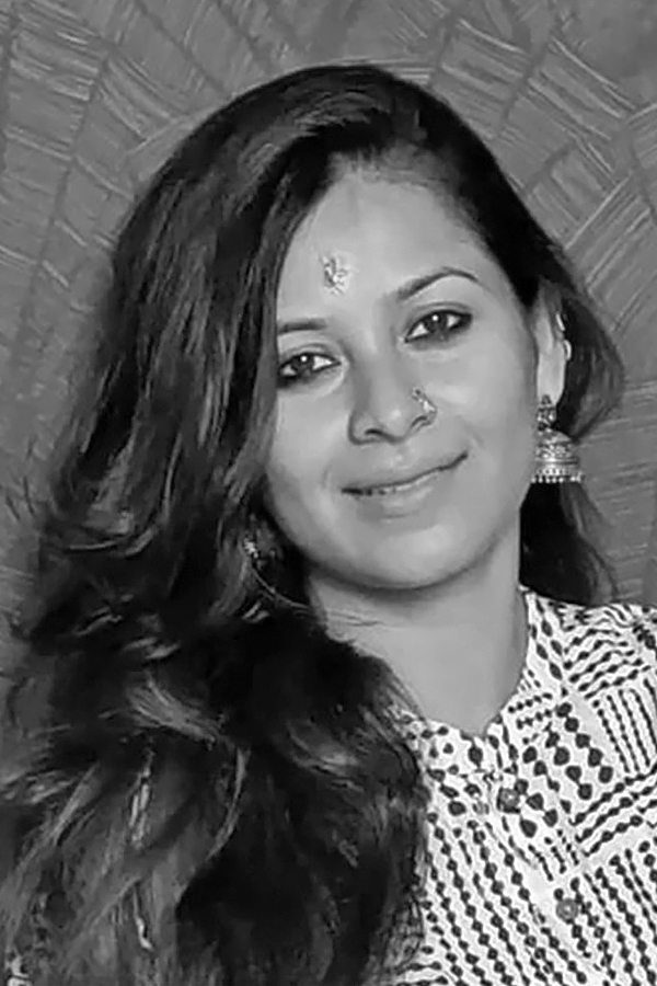 Angira in close portrait, smiling, her hair loose over one shoulder, wearing jhumka earrings and a patterned collar, a woven backdrop behind her. Black and white.