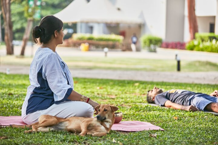 A facilitator seated on a mat on the lawn beside a participant lying down, a dog resting nearby