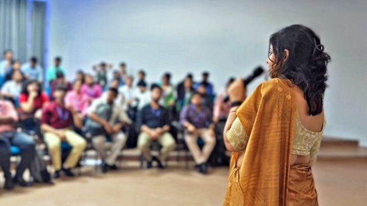 A facilitator with a microphone, seen from behind, facing rows of seated students