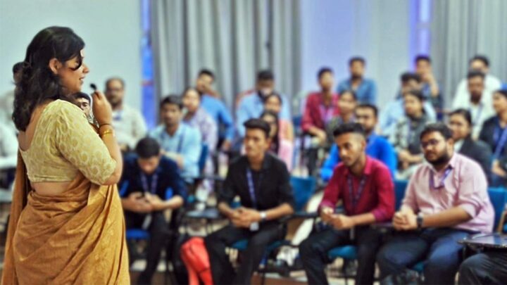A facilitator speaking into a microphone at the front of a lecture hall, students watching from tiered seating