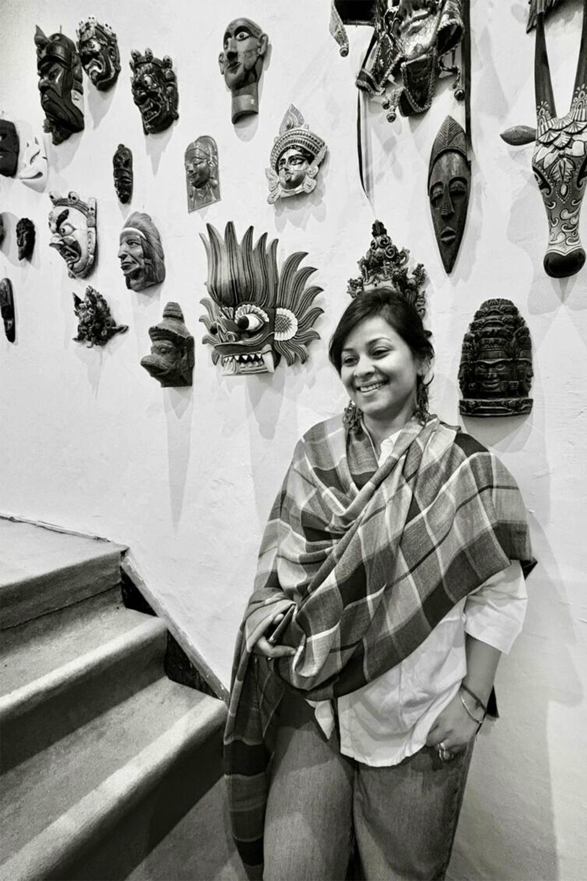 Angira Chakravorty Dasgupta standing beside a stairwell against a wall of carved and painted masks, in a black and white portrait.