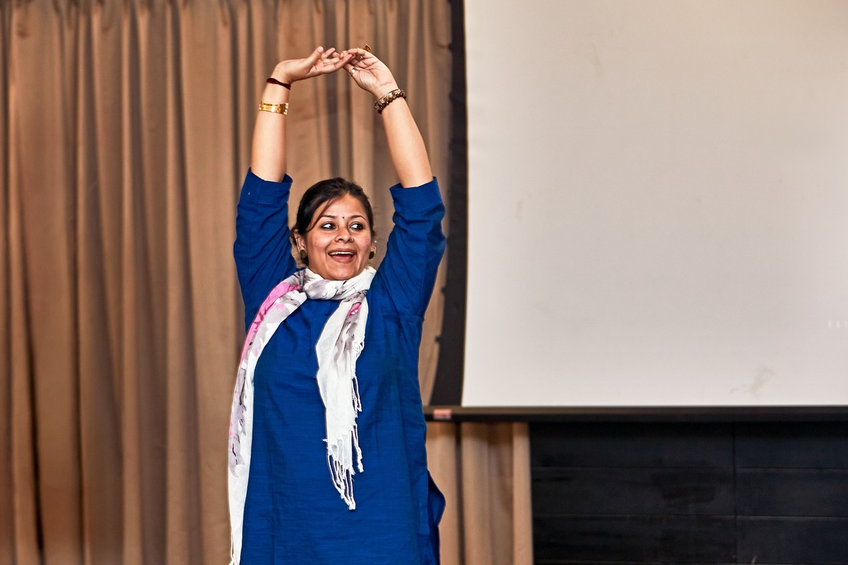 Angira Chakravorty Dasgupta on stage in a blue kurta, arms raised overhead with hands meeting, mid-movement during a corporate session.