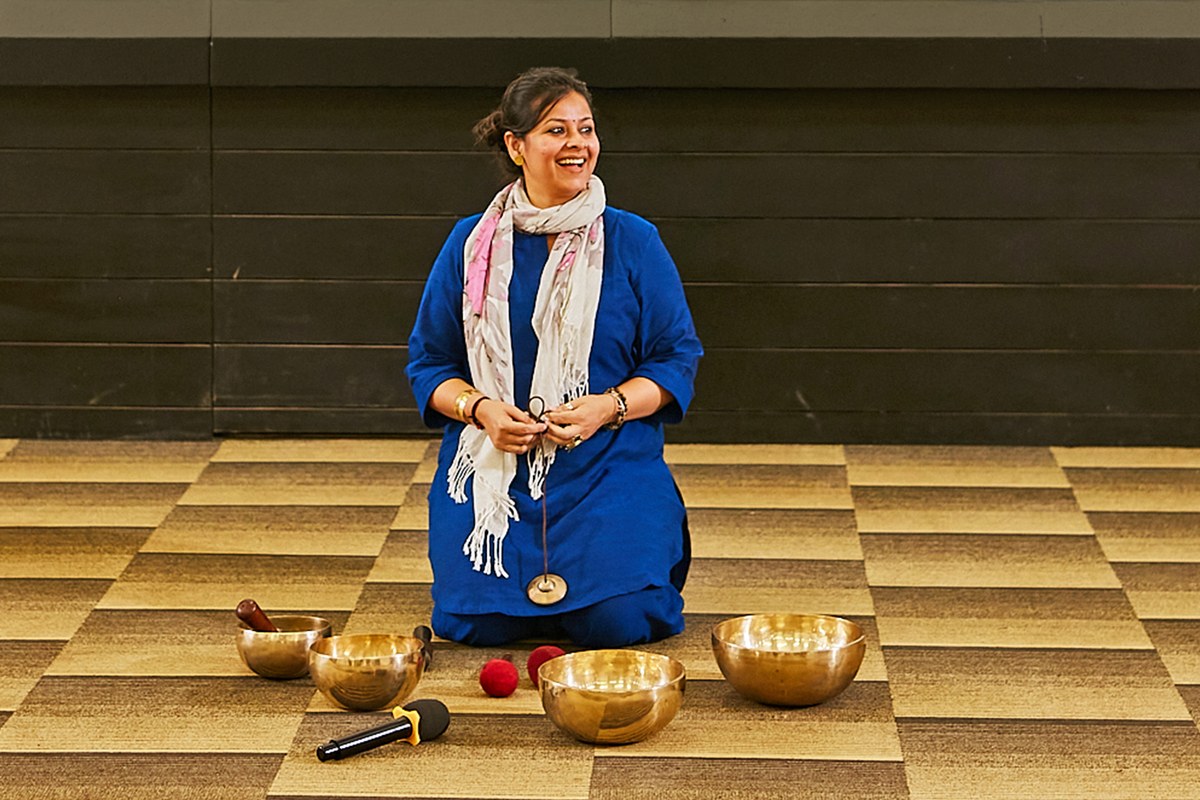 Angira Chakravorty Dasgupta kneeling on a chequered floor with four brass singing bowls and mallets set out in front of her before a sound therapy session.