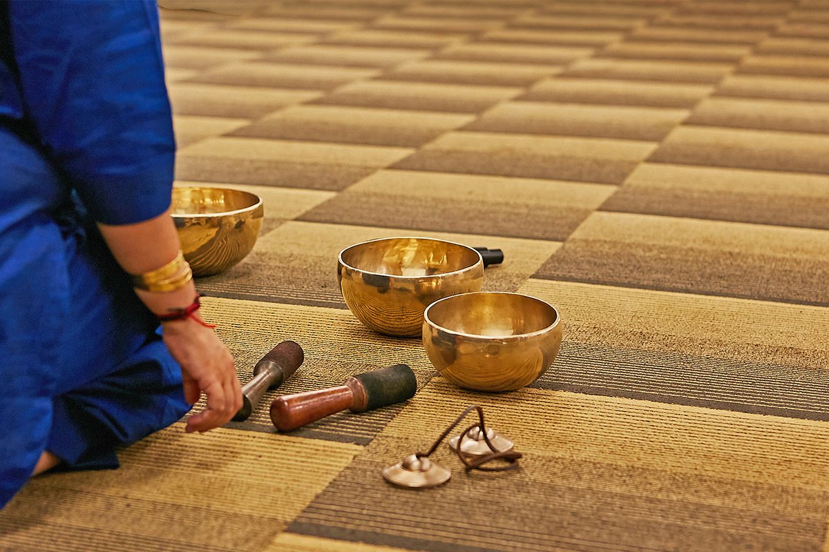 A hand holding a mallet above three brass singing bowls set out on a woven mat, before a sound therapy session.