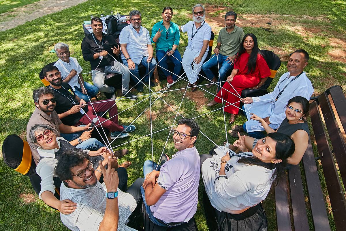 A group seated in a wide circle on the grass, holding a web of string stretched between them, photographed from above.