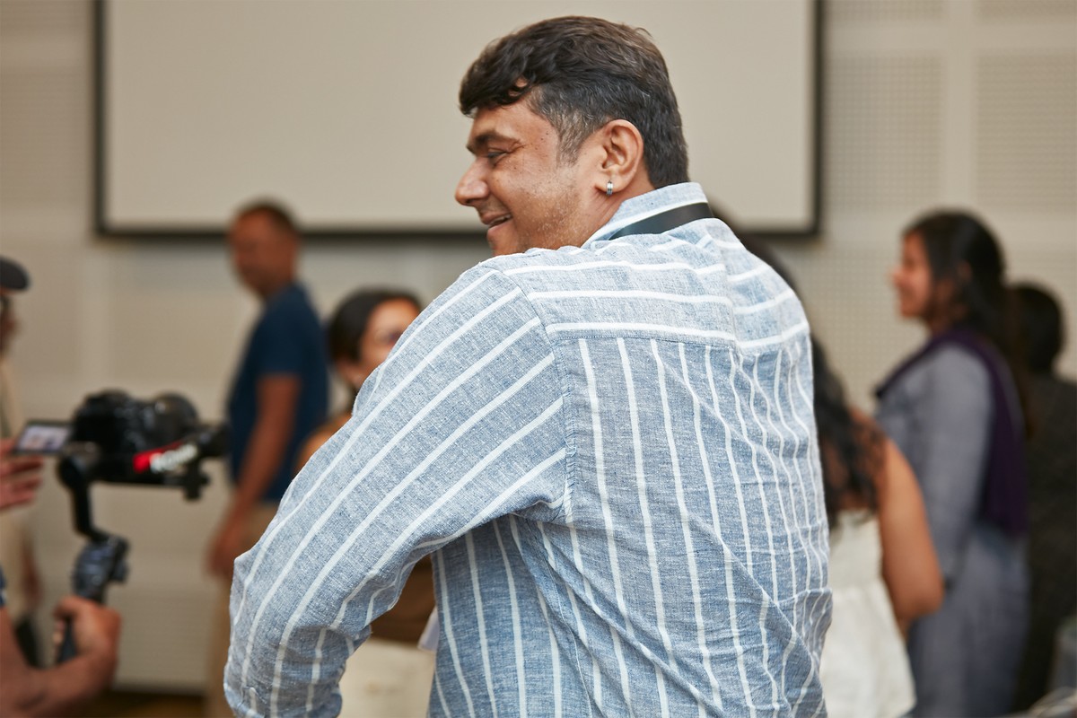 A participant in the foreground of an indoor corporate session, with colleagues and a camera on a tripod behind.