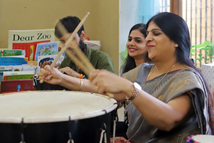 A participant in a grey and gold sari drumming with sticks, children's picture books on a shelf behind her