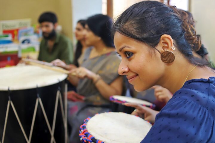 Close profile of a participant leaning over a djembe, smiling, a row of drummers behind her