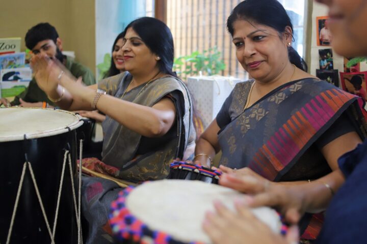 Two participants in saris drumming together, one striking with sticks, the other with an open palm