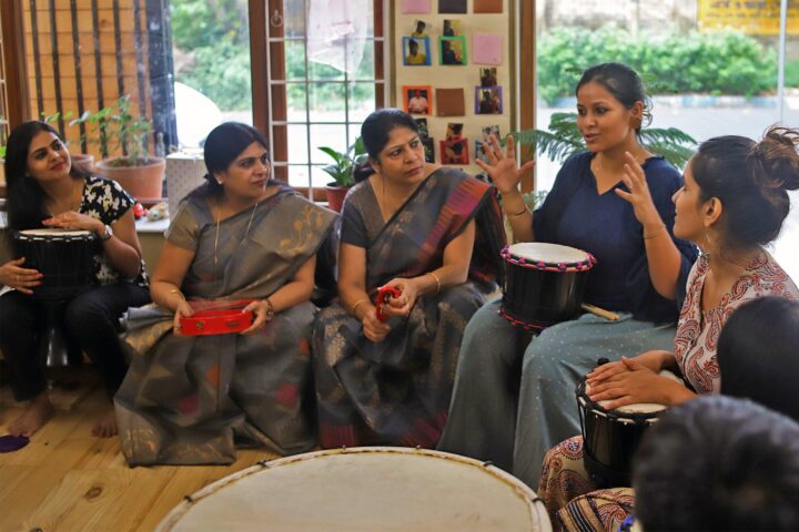 The facilitator explaining with both hands raised, seated among a circle of participants holding drums and a tambourine