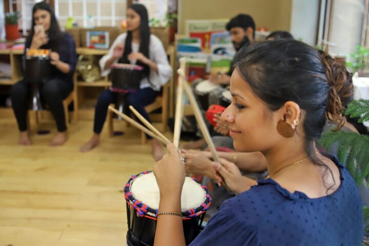 A participant holding drumsticks above a djembe, others seated on low stools across the wooden floor