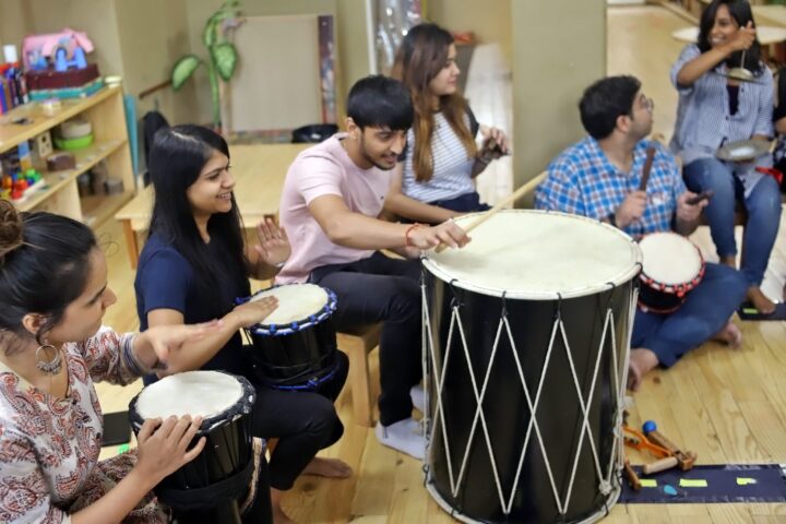 A mixed group seated around a large dhol, drumming and clapping together in a bright playroom