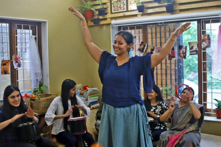 The facilitator standing with both arms raised, the seated circle clapping and drumming around her