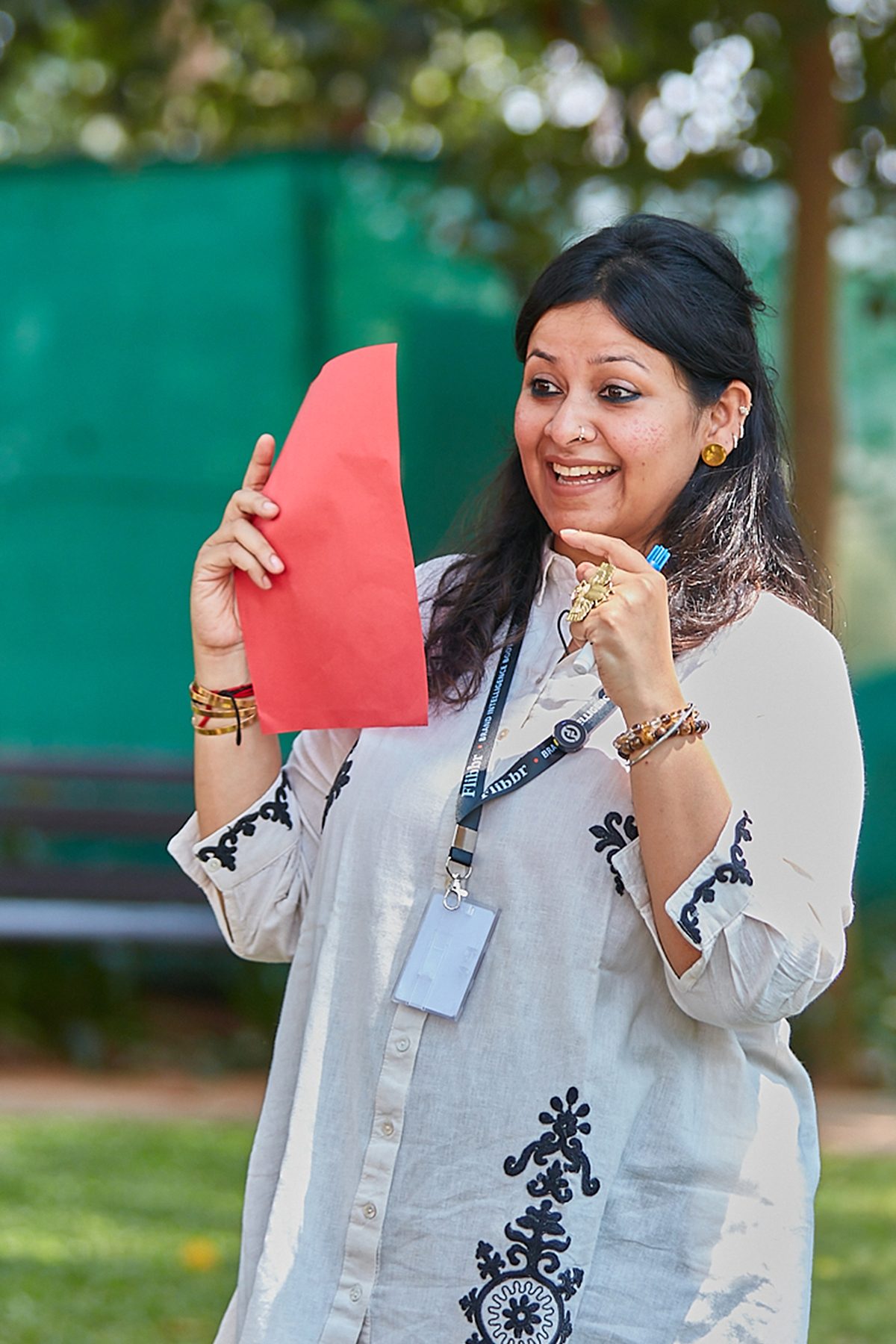 Angira Chakravorty Dasgupta outdoors at a workshop, holding up a coral card and smiling as she speaks, wearing a pale embroidered kurta and a lanyard.
