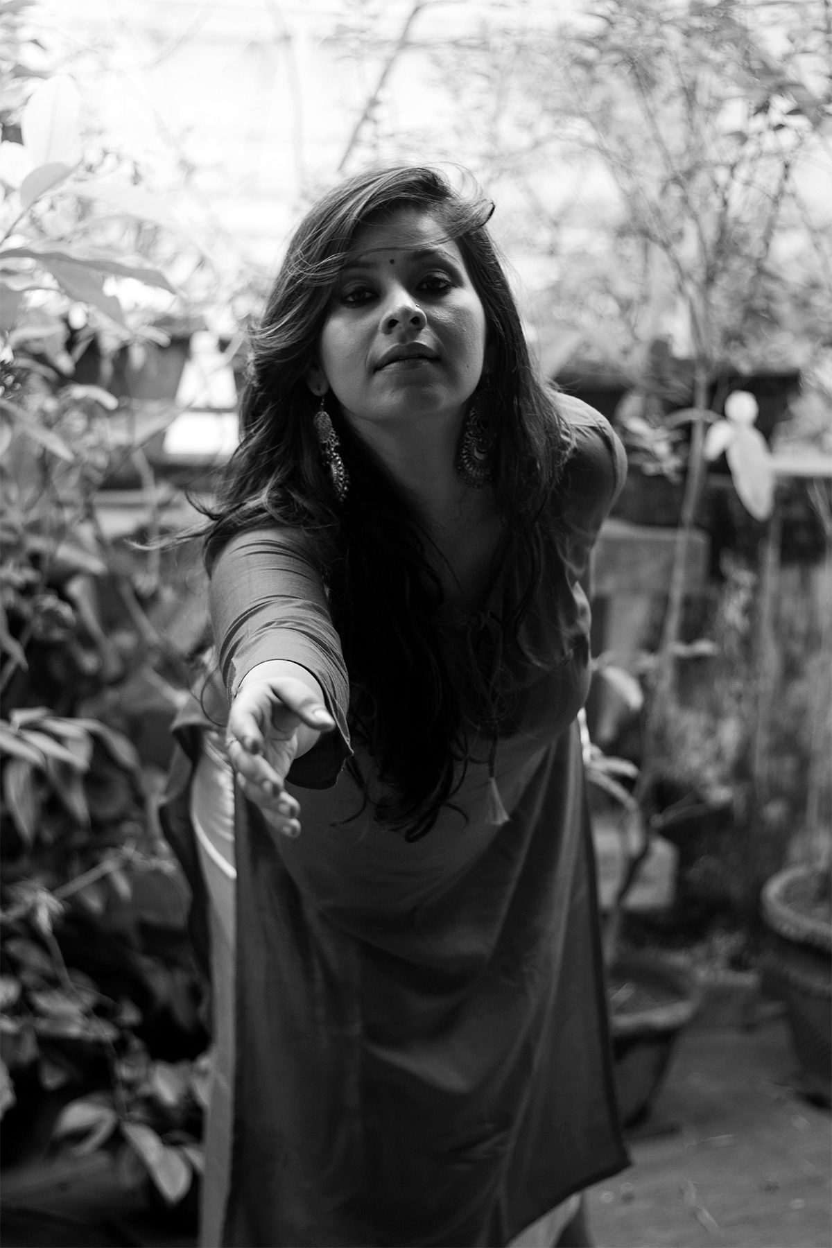 Angira Chakravorty Dasgupta outdoors among plants, one hand extended towards the camera, wearing a dark sleeveless kurta and filigree jhumka earrings. Black and white.