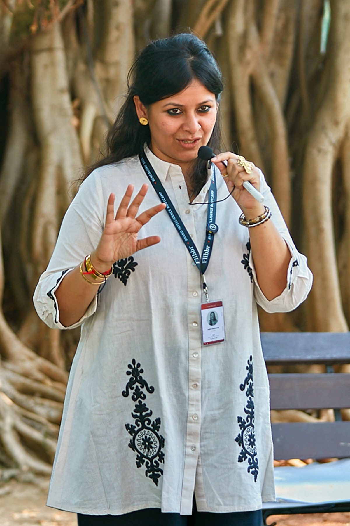 Angira Chakravorty Dasgupta speaking into a microphone beneath a banyan tree, one hand raised mid-gesture, wearing a white embroidered kurta and a lanyard. She runs SVA flo teacher wellbeing programmes for schools, colleges and universities.