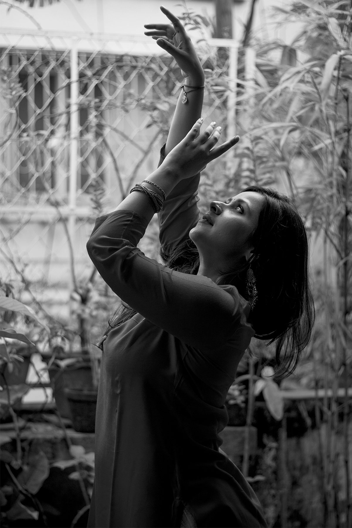 Angira Chakravorty Dasgupta outdoors among plants, both arms reaching upward, her face turned up, mid-movement.