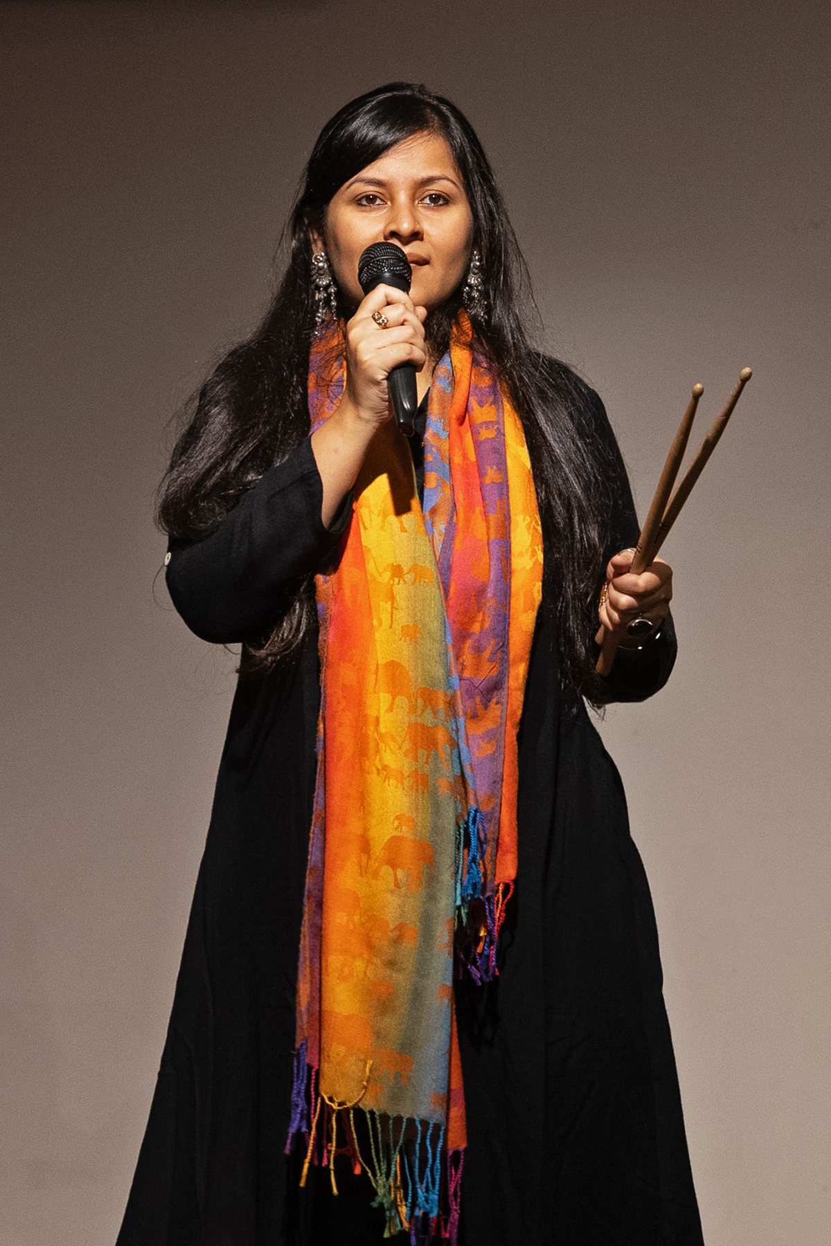 Angira Chakravorty Dasgupta on stage in a black kurta and bright striped scarf, speaking into a microphone with drumsticks in her other hand.