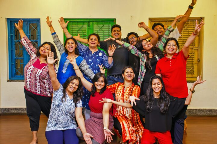 The whole group with arms raised in front of the studio's blue and green shuttered windows
