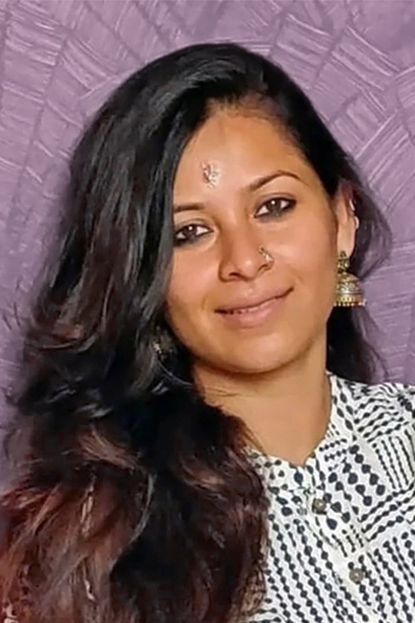 Angira in close portrait, smiling, her hair loose over one shoulder, wearing jhumka earrings and a patterned collar, a woven backdrop behind her.