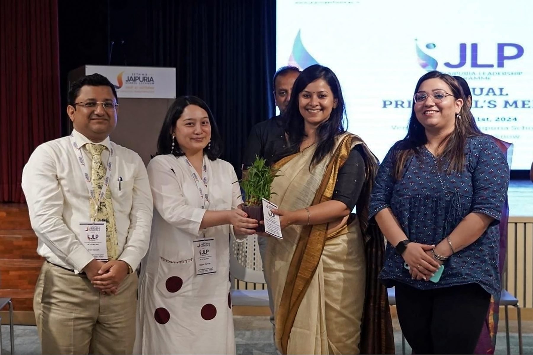 Angira on stage being presented with a potted plant, standing with three others in front of a Jaipuria Leadership Programme slide.