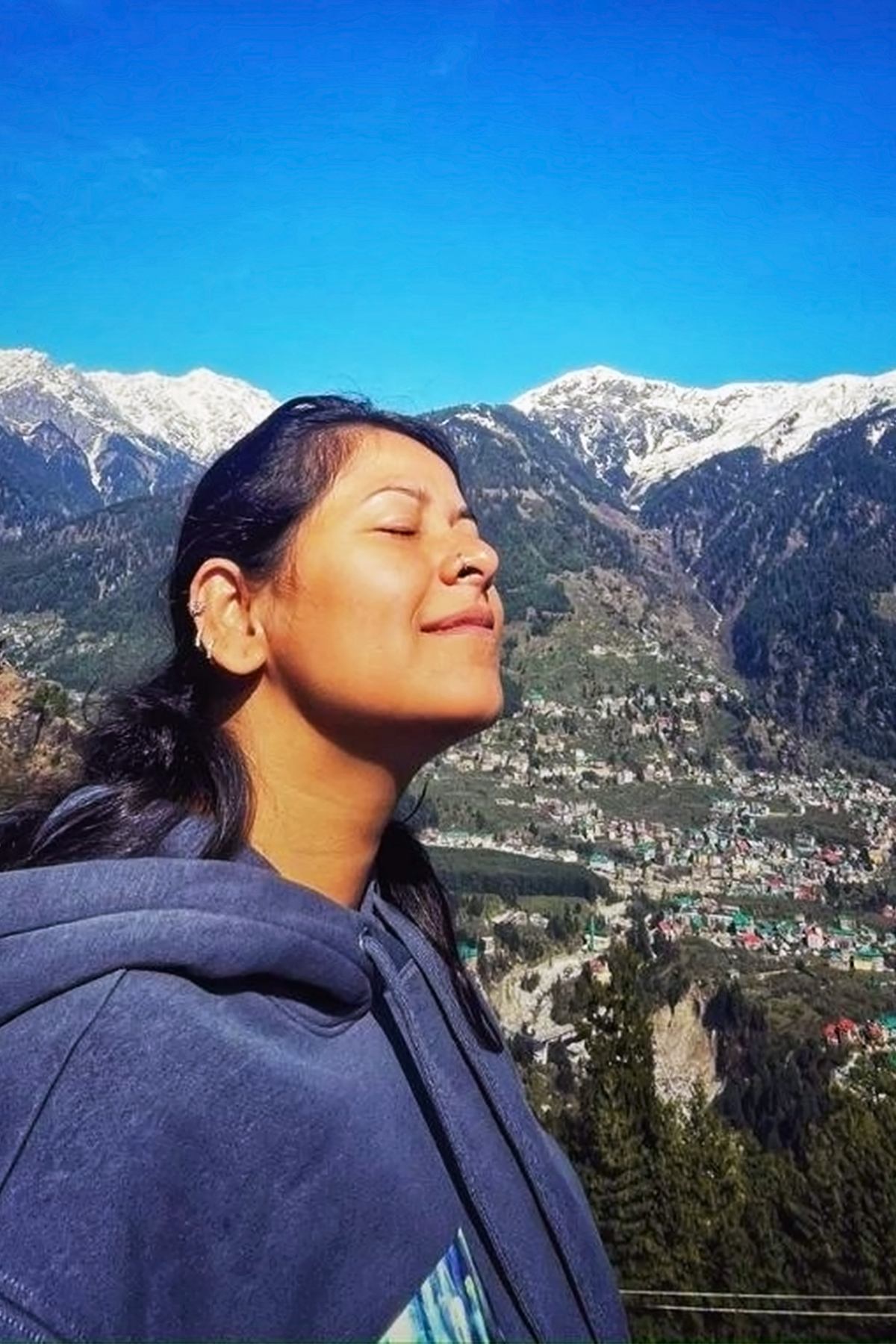Angira outdoors in the mountains, eyes closed and face turned up to the sun, snow-capped peaks and a valley town behind her.