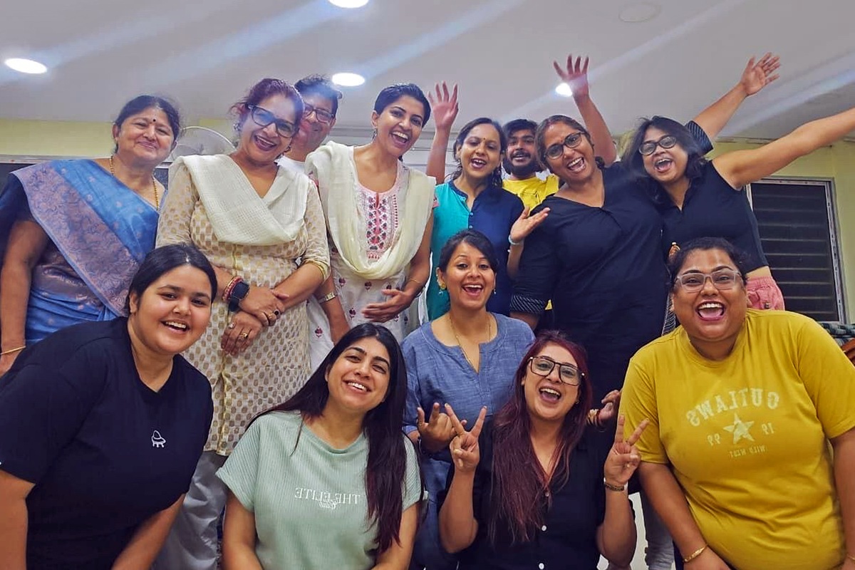 A workshop group laughing together indoors under ceiling lights, several with arms flung up and the front row making victory signs.